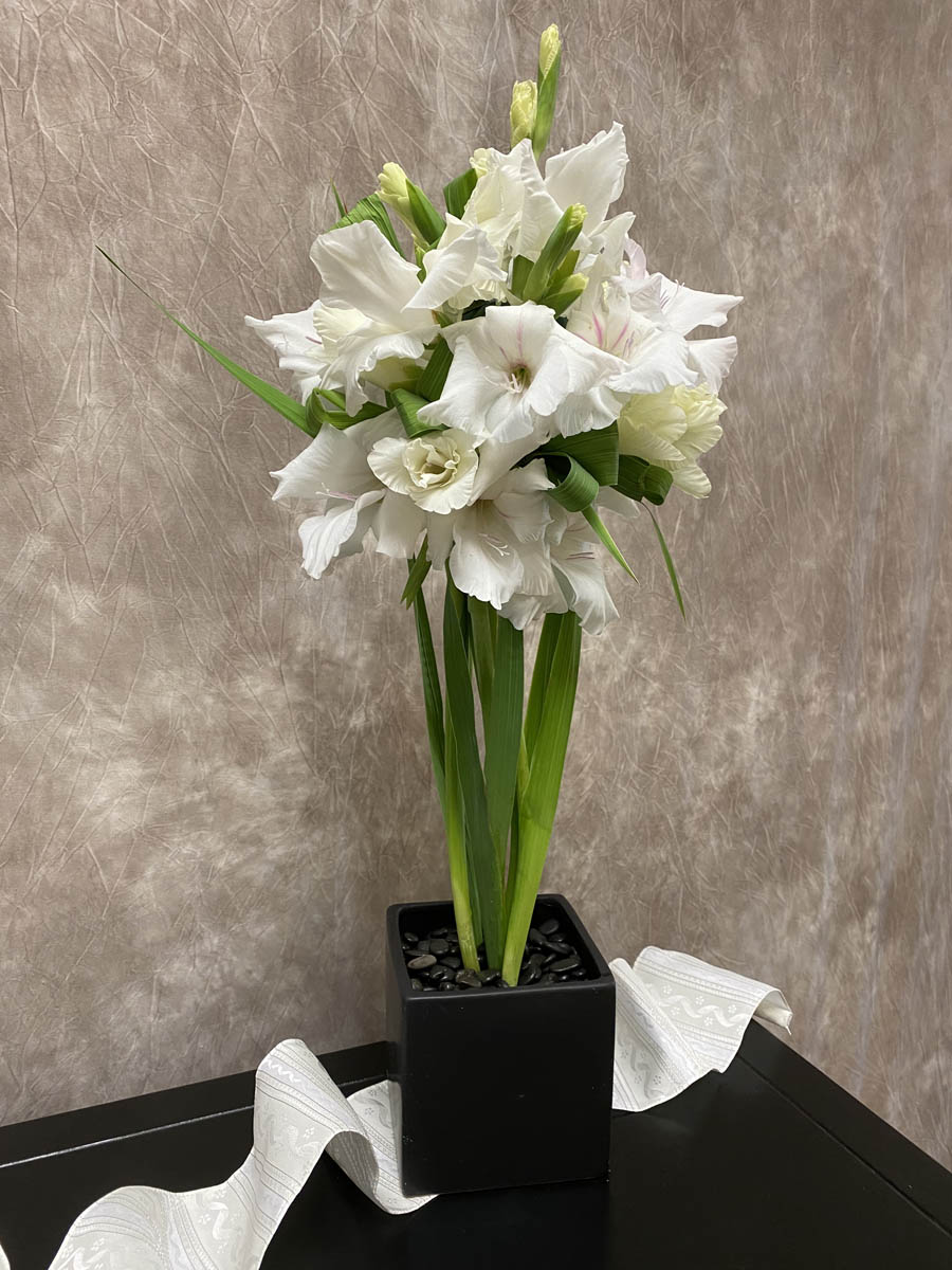 Figure 12 Finished DesignA beautiful arrangement of five white gladioli stems in a cubed container with decorative stones is displayed on a table with a decorative runner underneath.
